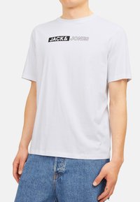 White cotton t-shirt featuring a black graphic label with "JACK&JONES" text. Short sleeves, crew neck, and relaxed fit. Casual style.