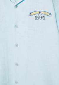 Chemise bleu clair à boutons avec une broderie indiquant "AMALFI COAST 1991" en jaune et bleu marine sur la partie gauche de la poitrine.