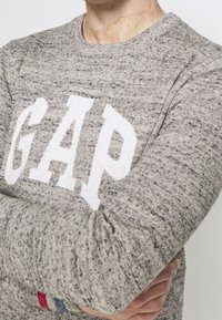 Szary dzianinowy sweter z dużym białym logo "GAP" na przodzie. Prążkowany okrągły dekolt i subtelna faktura tkaniny.