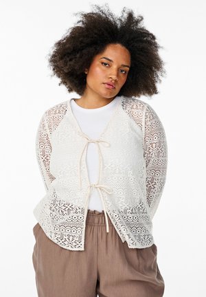 Spitzenstrickjacke in Off-White mit einer Bindefront, die ein florales Muster und eine halbtransparente Textur aufweist, kombiniert mit einem weißen Top und braunen Hosen.