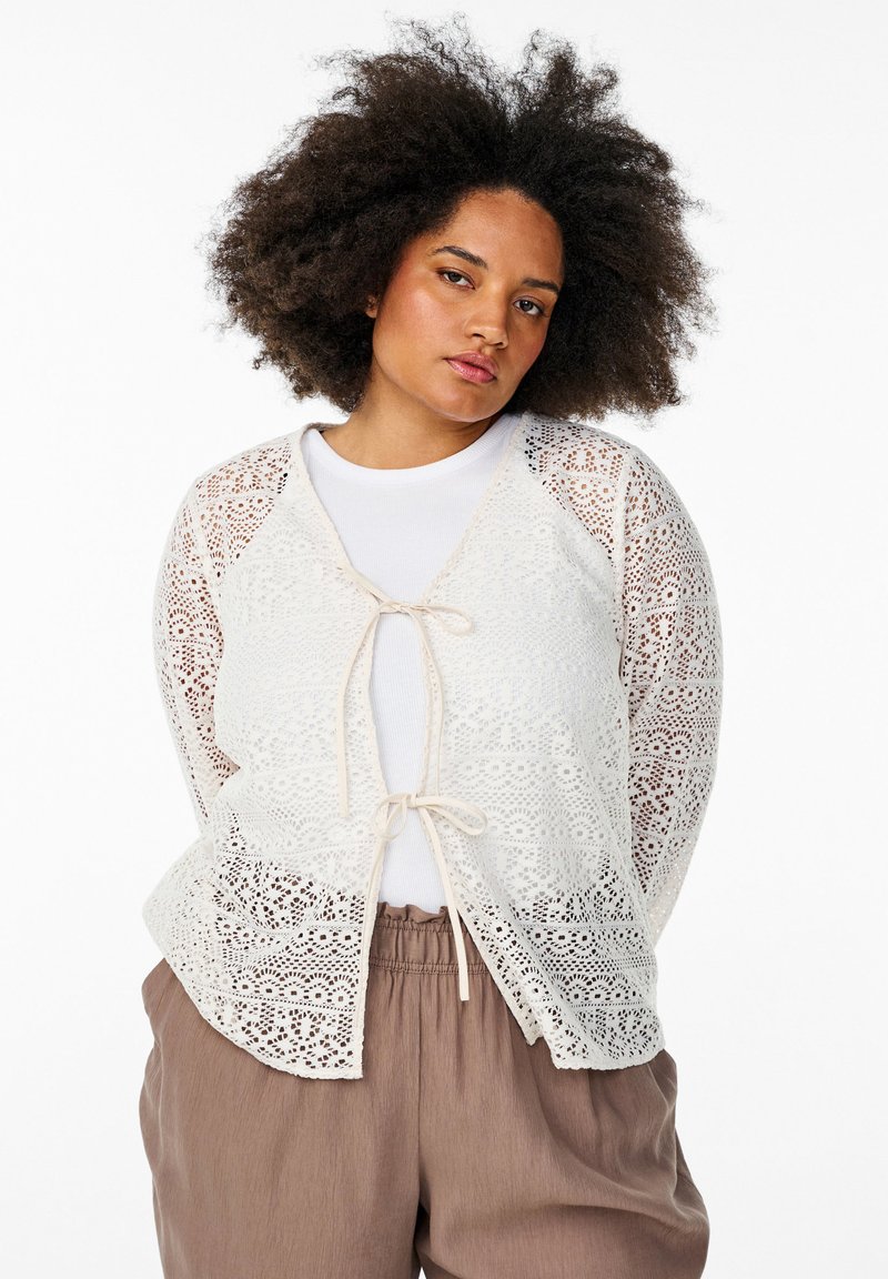 Cardigan en dentelle blanc cassé avec un devant noué, présentant un motif floral et une texture semi-transparente, assorti à un haut blanc et un pantalon marron.