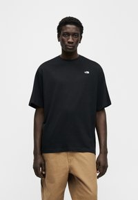 Νέος άνδρας που φοράει μαύρο t-shirt The North Face και μπεζ παντελόνι, στέκεται μπροστά σε ένα απλό φωτεινό γκρι φόντο.