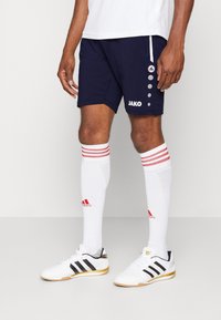 Pantaloni sportivi blu navy con logo JAKO, abbinati a calze bianche al ginocchio con strisce rosse e scarpe Adidas in bianco e nero.