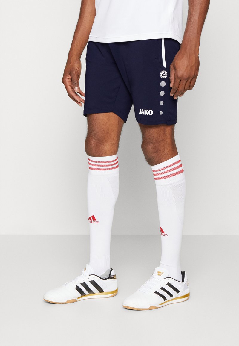Pantaloni sportivi blu navy con logo JAKO, abbinati a calze bianche al ginocchio con strisce rosse e scarpe Adidas in bianco e nero.