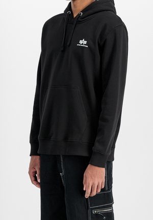 Osoba nosząca czarną bluzę z kapturem Alpha Industries z przednią kieszenią oraz czarne spodnie typu cargo z białym przeszyciem i kieszenią na zamek.