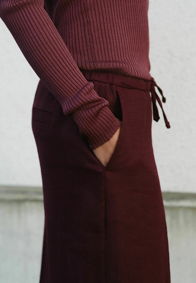 Personne portant un haut bordeaux côtelé à manches longues avec une main dans la poche d'un pantalon bordeaux assorti à cordon de serrage.