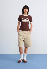 Brun t-shirt med vita detaljer och logotyp, beige shorts, vita strumpor och flerfärgade sneakers. Modellen står framför en blå bakgrund.
