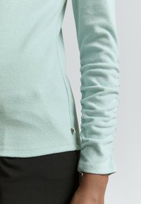 Jasnoniebieska koszula z długim rękawem z zaszewkami na rękawach, noszona z czarnymi spodniami, z naciskiem na bok i dolną część ramienia.