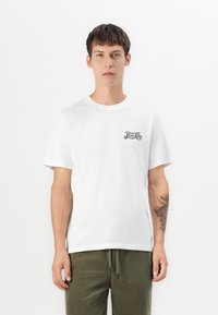 Young man wearing white Pepsi-Cola logo t-shirt and green drawstring pants, with tattoo on his left forearm, standing against white background.
