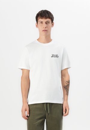 Young man wearing white Pepsi-Cola logo t-shirt and green drawstring pants, with tattoo on his left forearm, standing against white background.