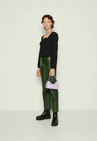 Fekete, hosszú ujjú felső, zöld bőr nadrág, fekete, vastag talpú csizmák, egy kisméretű levendula táskával ezüst részletekkel, semleges háttér előtt pózol.