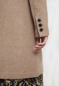 Manica di un cappotto di lana beige con tre bottoni neri e una mano rilassata di lato, tessuto a fantasia visibile nella parte inferiore.