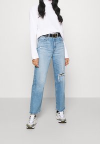 Hellblaue, ausgewaschene Jeans in lässiger Passform, kombiniert mit einem weißen, gerippten Rollkragenpullover und einem schwarzen Gürtel. Trägt dazu graue und weiße Sneakers.