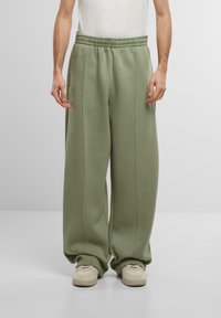 Pantalons de survêtement oversized vert clair avec une taille élastique et des coutures latérales, associés à des baskets beiges. Texture lisse, design décontracté.