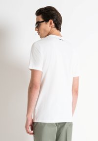 Camiseta de algodón blanca de manga corta, con un cuello redondo y un pequeño logo negro en la parte superior de la espalda. Llevada con pantalones verdes.