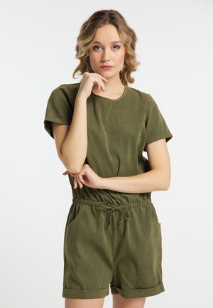 Junge Frau mit welligem blondem Haar trägt einen olivgrünen Kurzarm-Overall und steht mit der Hand am Kinn vor einem schlichten, hellen Hintergrund.