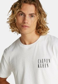 Camiseta de algodón blanca con texto negro "CALVIN KLEIN" en la parte izquierda del pecho, cuello redondo y mangas cortas con una textura suave.