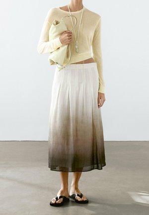 Personne portant un pull court beige clair, une jupe plissée en dégradé, des sandales noires, tenant un sac beige plié et portant plusieurs colliers longs superposés.