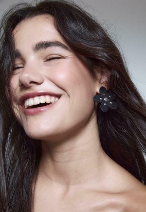 Orecchini a forma di fiore neri con accenti argentati, realizzati in materiale testurizzato, indossati da una persona con capelli scuri e un'espressione sorridente.