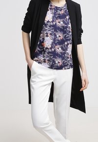 Cardigan lungo nero, blusa floreale blu navy e pantaloni bianchi su misura. La blusa presenta motivi floreali multicolore; texture del tessuto liscia.