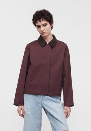 Person med kort krøllet hår iført en brun jakke med mørkraver og lyseblå revnede jeans, stående foran en ensfarvet baggrund.