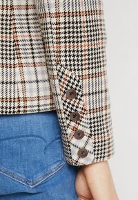 Manche de blazer à carreaux en tons crème, noir et orange, avec des boutons marron et un motif pied-de-poule. Jean avec détail de poche courbée visible.