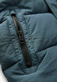 Veste isolante en tissu bleu foncé avec une fermeture éclair noire bien visible. Le matériau présente un motif texturé subtil et une apparence ondulée.