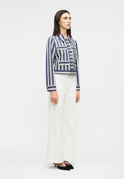 Γυναίκα στέκεται φορώντας λευκό παντελόνι με φαρδιά πόδια, σακάκι με κάθετες ρίγες σε σκούρο μπλε και λευκό με κουμπιά, και μαύρες ψηλοτάκουνες μυτερές γόβες, μπροστά σε απλό φόντο.