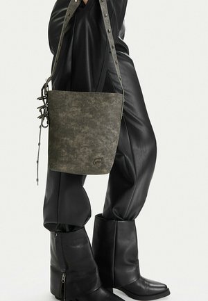 Bolso tipo cubo texturizado de color verde oliva con correa ajustable, llevado sobre pantalones de cuero negros y botas negras de cuero con cremallera.