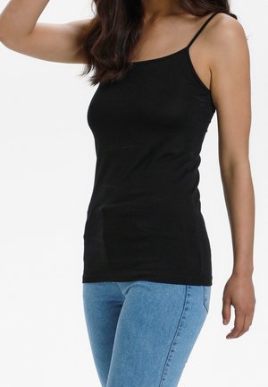 Femme portant un débardeur noir à fines bretelles et un jean bleu clair, posant avec un bras levé devant un fond uni.