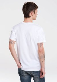 Camiseta de algodón blanca con mangas cortas, diseño de cuello redondo y textura suave. Lleva puesto un jean azul; tatuajes visibles en el brazo izquierdo.