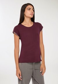 Kobieta stojąca, ubrana w bordową bluzkę z krótkim rękawem z perłowym wykończeniem oraz szare spodnie, lekko zwrócona w prawo na jednolitym tle.