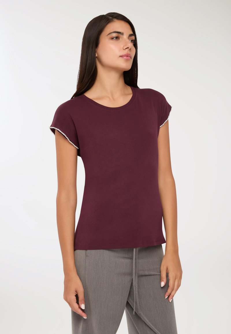 Kobieta stojąca, ubrana w bordową bluzkę z krótkim rękawem z perłowym wykończeniem oraz szare spodnie, lekko zwrócona w prawo na jednolitym tle.