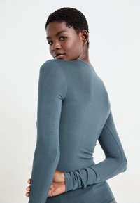 Haut à manches longues en tissu doux et bleu canard profond, avec un design ajusté et une texture lisse. Présente un col rond et des manches fines.
