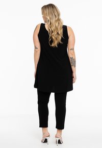 Schwarze ärmellose Tunika mit asymmetrischem Saum, kombiniert mit schwarzen Leggings. Das Modell trägt silberne Absätze. Glattes Gewebe mit lockerer Passform.