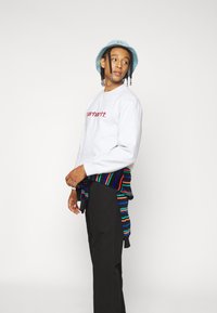 Grauer Sweatshirt mit rotem "Carhartt"-Logo, kombiniert mit schwarzen Hosen und einem mehrfarbigen, gestreiften Pullover, der um die Taille gebunden ist. Blauer Bucket Hat.