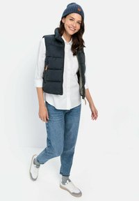 Gilet rembourré marine porté sur une chemise blanche, associé à un jean bleu et des baskets blanches. Le gilet a un col montant et une fermeture éclair, avec un accent en cuir.