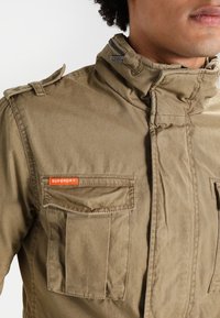 Detailní záběr osoby v khaki bundě s vysokým límcem a kapsami na přední straně, přičemž jedna z nich je označena nápisem "Superdry" na hrudi.