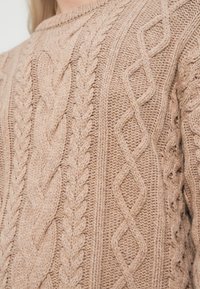 Beige cable-knit sweater featuring various textured patterns, including braids and diamonds, with a classic crew neckline.