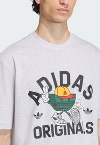 Sivé bavlnené tričko s farebnou karikatúrnou grafikou postavy v žltej čiapke, ktorá ukazuje znak mieru, text "ADIDAS ORIGINALS."