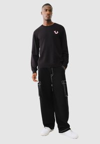 Schwarzer Pullover mit Rundhalsausschnitt und pinkem Logo, kombiniert mit schwarzen Cargo-Hosen mit weißen Nähten und seitlichen Taschen, getragen mit weißen Sneakern.
