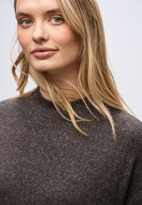 Maglione lavorato a maglia grigio scuro con collo alto, morbida texture e un sottile motivo mélange, indossato da una persona con lunghi capelli biondi.