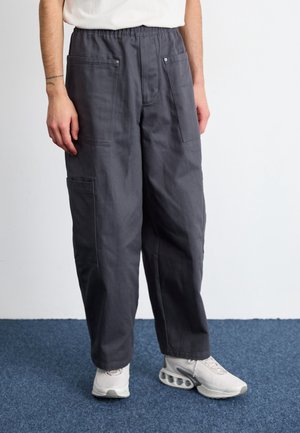 Homme portant un pantalon cargo ample gris foncé avec plusieurs poches et des baskets blanches, debout sur un tapis bleu contre un mur blanc.