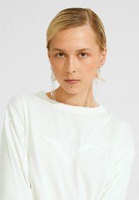 Vrouw met naar achteren gekamd blond haar, die lange zilveren kettingoorbellen en een witte top draagt, en recht in de camera kijkt.