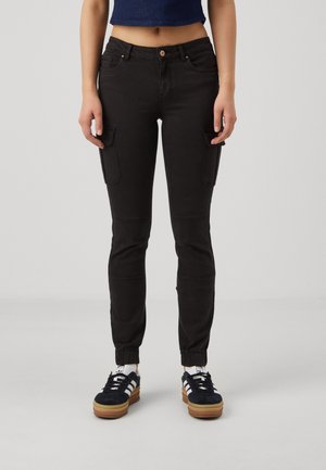 Kvinde iført sorte cargobukser med sidelommer og elastiske manchetter, kombineret med sort-hvide sneakers, stående mod en ensfarvet baggrund.