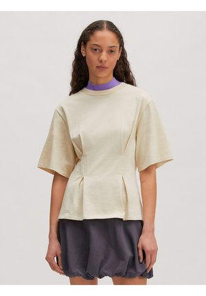 Frau mit langen lockigen Haaren, die ein cremefarbenes Kurzarmshirt über einem violetten Rollkragenpullover und einem dunkelgrauen Ballonrock vor einem schlichten Hintergrund trägt.