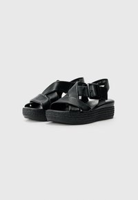 Sandales à plateforme noires avec des brides en cuir croisées et une boucle décorative. Elles disposent d'une semelle texturée noire et d'un talon plat.