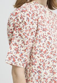 Blouse fleurie à manches courtes, avec un fond crème et des fleurs rouges, brunes et bleues. Tissu texturé et détails froncés aux épaules.
