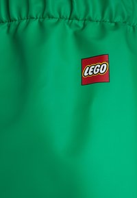 Zeleno blago z gladko teksturo, ki vsebuje rdeči Lego logotip. Material je rahlo sijoč in zložen na vrhu.