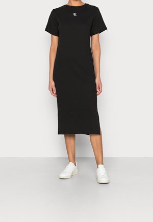 Personne portant une robe midi noire à manches courtes avec un petit logo blanc sur la poitrine et des baskets blanches, debout devant un fond clair uni.